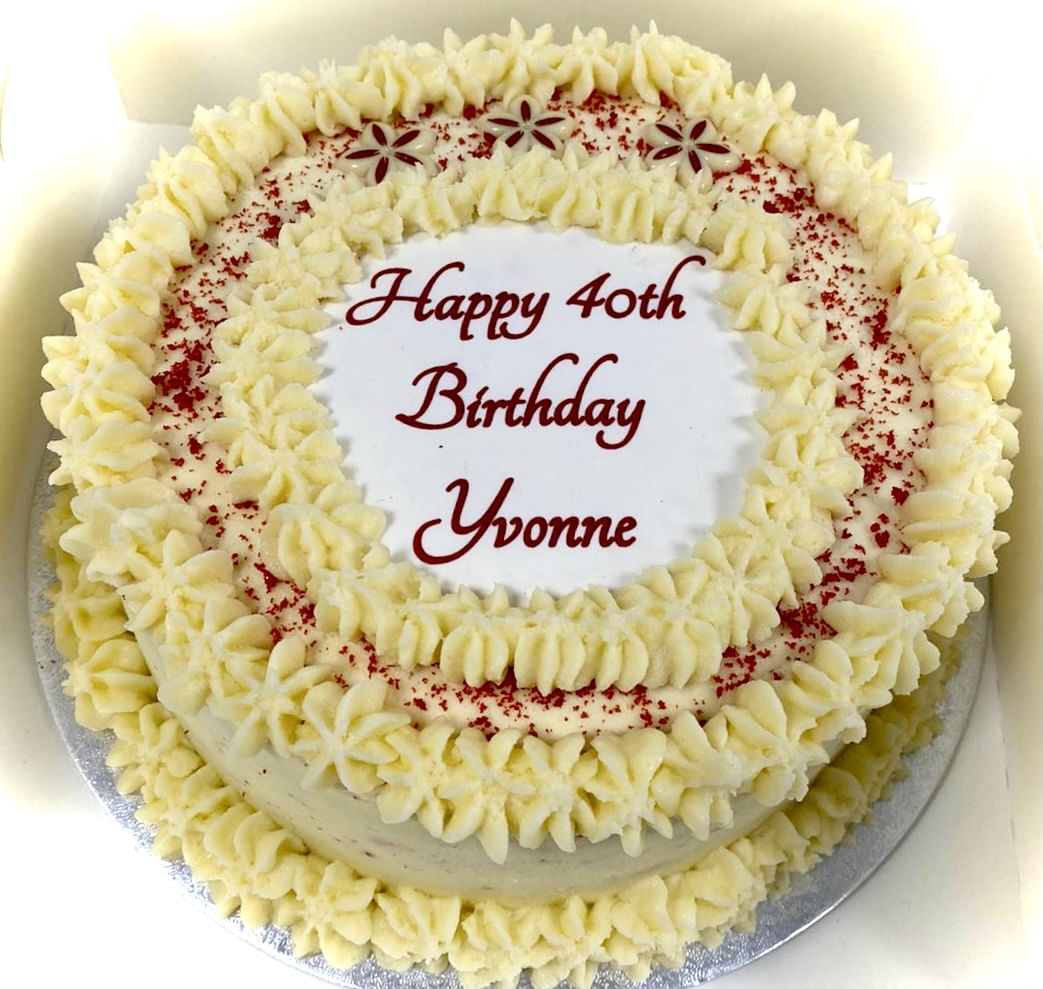 A special occasion round Red Velvet Cake decorated with white cream cheese frosting and red sprinkles. The top of the cake has a piped cream border and a message in the center that reads, 'Happy 40th Birthday Yvonne' in red. Small decorative flowers are also on the top.